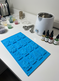 A blue shallow mold with 16 cavities, each depicting a different Christmas-themed design, such as Santa, reindeer, and festive greetings.